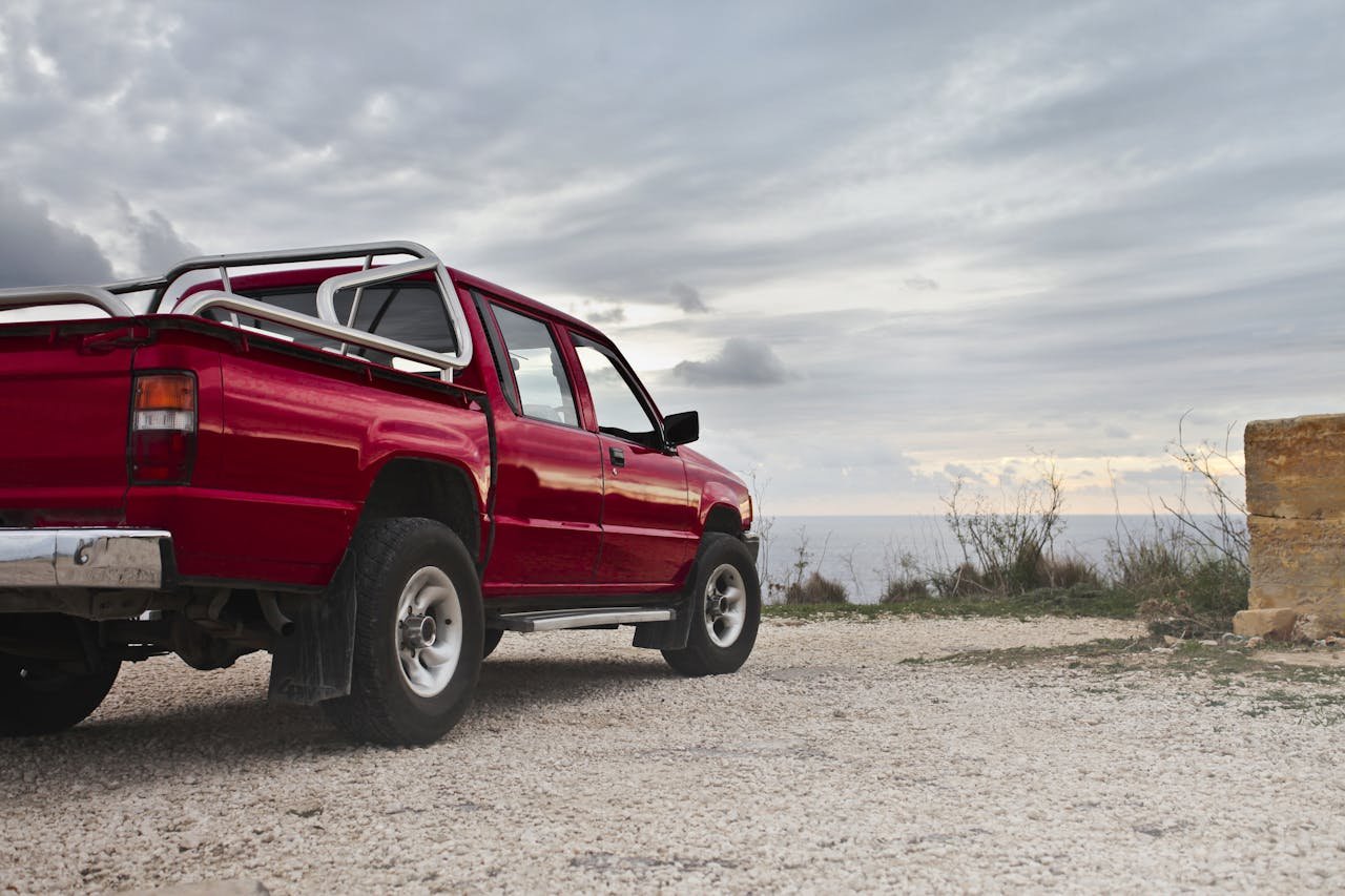 A red pickup truck parked by the rocky coastline under a cloudy sky in Mgarr, Malta.